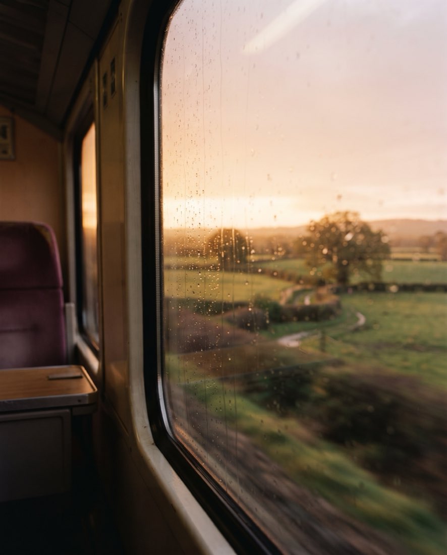 Countryside at golden hour seen through a rain-flecked train window from inside a carriage.